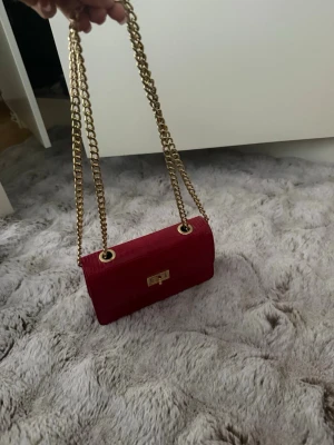 Vinröd axelremsväska med guldkedja - Snygg vinröd handväska med krokodilpräglat mönster och guldfärgad kedjerem. Väskan har ett elegant metallspänne framtill och en stilren, rektangulär form. Perfekt för dig som vill ha en trendig accessoar med lyxig känsla.