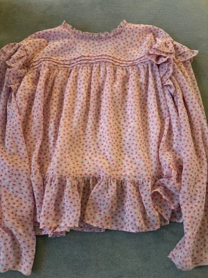 Rosa blommig volangblus - Söt rosa blus med små blommor i rött och vitt. Blusen har volangdetaljer vid axlar och nederkant samt långa, puffiga ärmar. Materialet är tunt och luftigt, perfekt för en romantisk look. Rund halsringning med liten volangkant upptill.