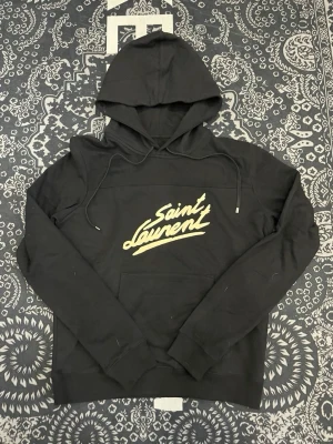 Svart Saint Laurent hoodie - Snygg svart hoodie från Saint Laurent med stor vit logga i graffiti-stil på bröstet. Klassisk känguruficka framtill, dragsko i huvan och ribbade muddar. Perfekt för dig som gillar streetwear och vill ha något stilrent men ändå statement. Mjuk bomullskänsla.