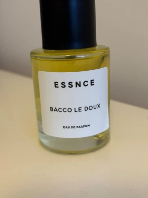 ESSNCE Bacco Le Doux Eau de Parfum - Modern parfym från ESSNCE med namnet Bacco Le Doux. Flaskan är cylindrisk i genomskinligt glas med svart lock och vit etikett med svart text. Innehållet är ljusgult och flaskan rymmer 50 ml Eau de Parfum. En stilren och minimalistisk doftflaska.