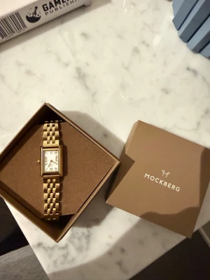 Guldfärgad klocka från Mockberg - Snygg guldfärgad klocka från Mockberg med rektangulär urtavla och klassisk länkarmband. Urtavlan är vit med romerska siffror och gulddetaljer. Kommer i originalask och extra länkar medföljer. Perfekt accessoar för en stilren look.