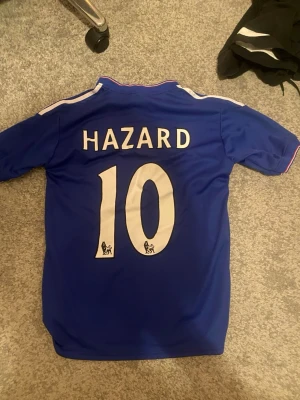 Chelsea Hazard matchtröja blå - Säljer en blå Chelsea FC matchtröja från Adidas med Hazard och nummer 10 på ryggen. Tröjan har vita detaljer, klubbmärke på bröstet och Yokohama Tyres som sponsortryck. Klassisk krage med knappar och tunnt, ventilerande material perfekt för fotboll.