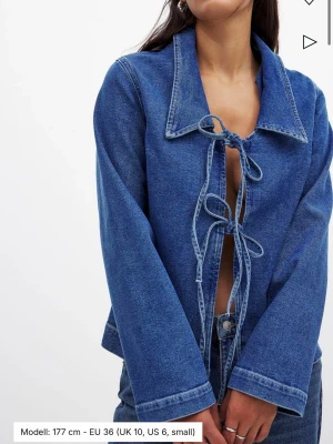 Blå jeansjacka med knytband NA-KD - Blå jeansjacka från NA-KD med bred krage och två knytband framtill. Jackan har raka ärmar och en avslappnad passform. Som ny i skicket, aldrig använd. 