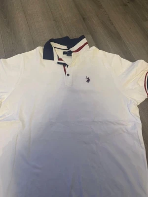 Vit piké från U.S. Polo Assn - Klassisk vit pikétröja från U.S. Polo Assn med broderad logga på bröstet. Tröjan har korta ärmar, krage i marinblått och detaljer i rött och vitt. Perfekt till jeans eller shorts för en clean och sportig look.