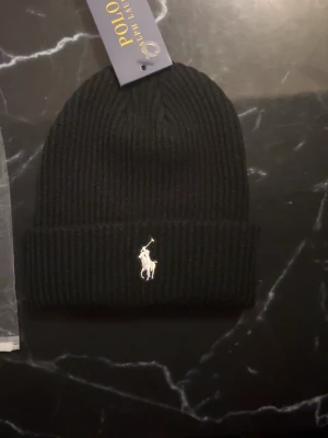 Svart ribbstickad mössa Polo Ralph Lauren aldrig använd helt svart bra kvalitet  - Svart ribbstickad mössa från Polo Ralph Lauren med klassisk uppvikt kant och broderad vit logga framtill. Snygg och enkel design som passar till det mesta. Perfekt accessoar för kyliga dagar.