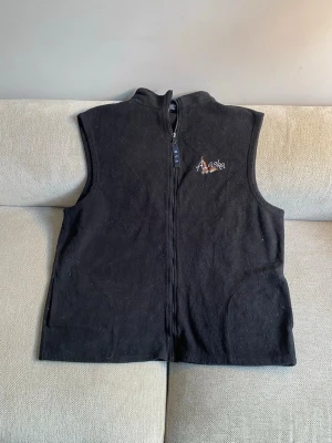 Svart fleeceväst - Svart fleeceväst med dragkedja framtill och broderad Alaska-logga på bröstet. Västen har två fickor på sidorna. Perfekt för lager-på-lager och enkel att matcha med olika outfits.