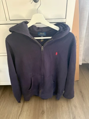 Mörkblå hoodie från Polo Ralph Lauren - Mörkblå hoodie med dragkedja från Polo Ralph Lauren. Klassisk design med huva, kängurufickor och den ikoniska röda logotypen broderad på bröstet. Tillverkad i mjukt material som är skönt att ha på sig. Perfekt för en avslappnad och stilren look. Lite smutsig för den har legat i en kartong 