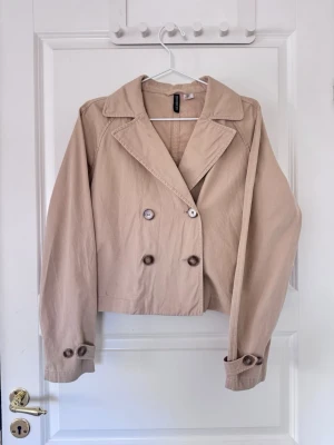 Kort beige trenchcoat  - Stilren kort trenchcoat i beige från H&M med dubbelknäppning framtill, klassisk krage och knappar vid ärmslut. Jackan har en avslappnad passform och är tillverkad i ett lätt bomullsmaterial.🌸🌸💕