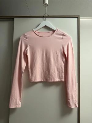 Ljusrosa croppad långärmad topp - Säljer en ljusrosa croppad långärmad topp i mjuk bomull. Enkel och stilren design med rund halsringning och tight passform. Perfekt att matcha med höga jeans eller kjol.