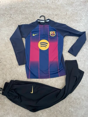 Barça träningsset Nike långärmad - Säljer ett FC Barcelona träningsset från Nike med långärmad tröja i blått och vinrött med klubbmärke och sponsorlogga på bröstet. Matchande svarta träningsbyxor med gul Nike-logga. Perfekt för fotbollsträning eller chill.