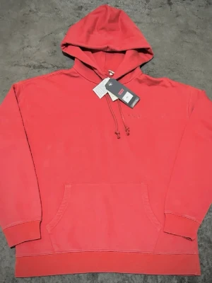 Röd hoodie från Levi's med huva - Snygg röd hoodie från Levi's med klassisk känguruficka och justerbar huva med dragsko. Hoodien har ribbade muddar vid ärmslut och nederkant samt diskret Levi's-logga på bröstet. Perfekt för en avslappnad och trendig look.