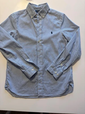  Polo Ralph Lauren - Klassisk blårandig skjorta från Polo Ralph Lauren i slim fit-modell. Skjortan har button-down krage, långa ärmar och broderad logga på bröstet. Tillverkad i bomull och har vita knappar framtill och vid ärmsluten. Perfekt för en stilren look. Stl 14 sitter som xs skulle säga passar 160-170 lång. Pris kan diskuteras💯
