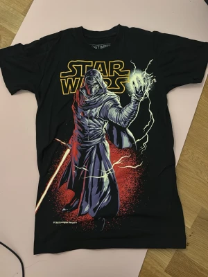 Star Wars svart t-shirt Kylo Ren - Säljer en svart Star Wars t-shirt med Kylo Ren-tryck i rött, vitt och gult. T-shirten har korta ärmar och en klassisk passform. Perfekt för dig som älskar Star Wars och vill ha en cool grafisk tee i garderoben. Storlek S men är ganska liten i passform.