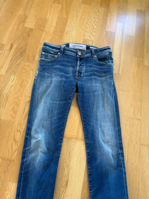 Blå jeans från Jacob Cohen - Säljer ett par snygga blå jeans från Jacob Cohen med klassisk femficksmodell och raka ben. tvättade detaljer för en cool, avslappnad look. Tillverkade i mjukt denimtyg med tydliga sömmar och knappgylf.OBS! lite hål på högra benet går att fixa!