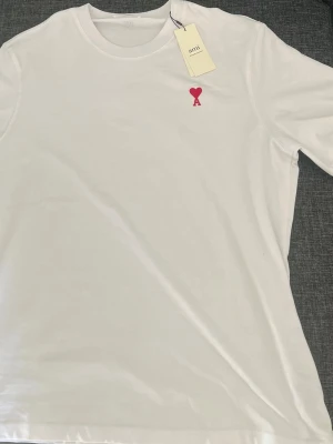 Vit t-shirt från Ami med rött hjärta - Säljer en stilren vit t-shirt från Ami Alexandre Mattiussi med det ikoniska röda hjärtemärket broderat på bröstet. Klassisk passform och rund halsringning. Perfekt basplagg för en clean look.
