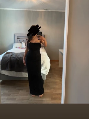 Oh Polly Palermo - Säljer en svart, glittrig långklänning med figurnära passform och axelbandslös design. Klänningen har en elegant slits bak och är perfekt för dig som vill sticka ut på festen. Materialet är stretchigt och skimrar snyggt i ljuset.