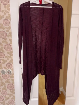 Vinröd lång kofta med öppen front - Säljer en vinröd lång kofta med öppen front och asymmetrisk nederkant. Koftan är tunn och har långa ärmar, perfekt att slänga över en topp för en avslappnad look. Materialet är lätt och något transparent, vilket ger en snygg layering-effekt.