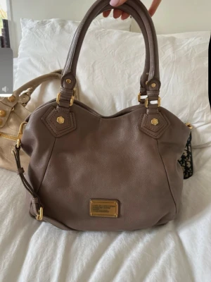 Brun handväska från Marc by Marc Jacobs - Snygg brun handväska i mjukt läder från Marc by Marc Jacobs. Väskan har dubbla handtag, guldfärgade metalldetaljer och en metallbricka framtill med logga. Klassisk och rymlig modell med avtagbar axelrem. Pris kan diskuteras 