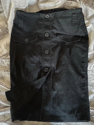 Svart knappkjol med fickdetaljer - Säljer en svart långkjol med stora knappar framtill och två dekorativa fickor med lock. Kjolen har en rak passform och slits framtill för extra rörelsefrihet. Perfekt till både sneakers och boots för en cool vardagslook.