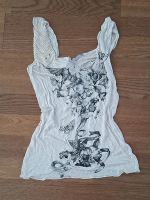 Beige linnetopp med blommönster - Snygg beige linnetopp med svart blommönste framtill. Ena axelbandet har dekorativa paljetter. Toppen är ärmlös och har en figurnära passform. 