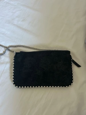 Svart kuvertväska med kedja - Snygg svart kuvertväska i mockaimitation med silverfärgade nitar längs kanterna och en cool silverkedja som axelrem. Väskan har dragkedja upptill och en stilren, rektangulär form. Perfekt accessoar för att lyfta din outfit.