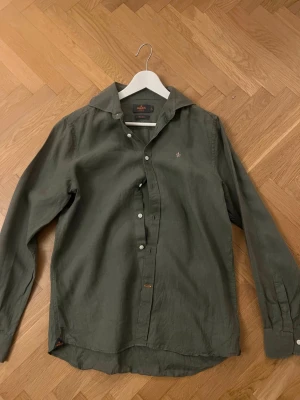 Olivgrön linneskjorta från Morris - Snygg olivgrön skjorta från Morris i 100% linne. Skjortan har klassisk krage, vita knappar och en liten broderad logga på bröstet. Storlek M, skriv för fler bilder 