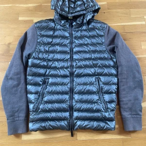 Grå pufferjacka med huva och stickade ärmar - Säljer en grå pufferjacka med glansig kropp och huva samt stickade, mörkgrå ärmar. Jackan har dragkedja framtill och två fickor med dragkedja. Perfekt för kyliga dagar.