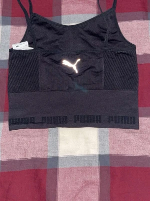 Svart sport-topp från Puma - Snygg svart sport-topp från Puma med smala axelband och elastisk nederkant med Puma-logga runtom. Materialet är stretchigt och andas, perfekt för träning. Reflekterande Puma-logo framtill och diskret mönster i tyget. Väldigt bekväm att ha på sig. Helt ny och oanvänd, men utan prislapp.