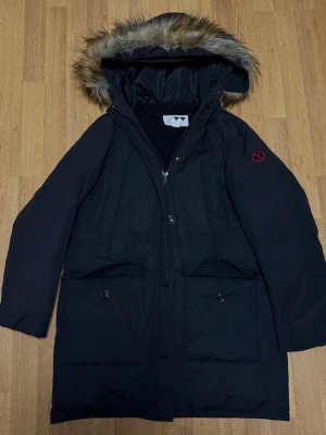 Svart parkas med fuskpäls och luva - Svart parkas med luva och fuskpäls, dragkedja och knappar framtill. Två stora fickor med lock och knapp. Fodrad med mjuk teddy på insidan upptill och slät polyester nedtill. Röd detalj på ärmen. Perfekt för kalla dagar. OBS 2 knappar saknas vid fästet av pälsen.