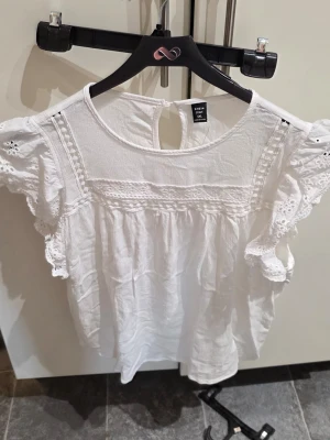 Vit blus med spetsdetaljer från SHEIN - Söt vit blus från SHEIN Curve med broderade spetsdetaljer och volang vid ärmarna. Blusen har rund halsringning, knapp i nacken och korta ärmar med hålmönster. Perfekt för en fräsch och somrig look.