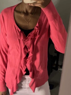 Rosa volangblus med öppen front - Trendig rosa blus med volangdetaljer längs den öppna framsidan. Blusen har rund halsringning, långa ärmar och ribbade muddar vid ärmslut och nederkant. Tillverkad i ett mjukt, stickat material som ger en bekväm och snygg passform.