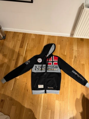 Svart hoodie Geographical Norway - Snygg svart hoodie från Geographical Norway med gråa muddar och huva. Framsidan har broderade och tryckta detaljer i vitt, rött och grått samt Norges flagga. Fickor framtill och dragkedja. Perfekt för dig som gillar streetwear och sportig stil.