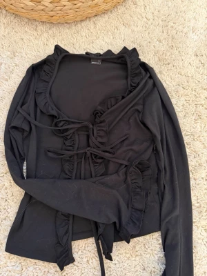 Svart omlottopp med volang från Gina Tricot - Säljer en svart omlottopp från Gina Tricot med långa ärmar och volangdetaljer längs kanterna. Toppen har knyt framtill och är tillverkad i ett mjukt material som känns skönt mot huden. Perfekt för dig som gillar trendiga detaljer och en feminin look.