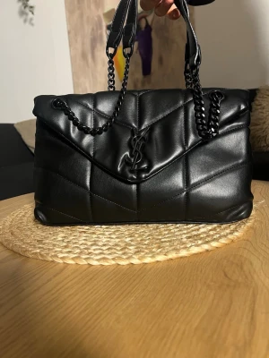 Svart quiltad handväska från YSL - Snygg svart  handväska från YSL med kedjehandtag och ikonisk YSL-logga framtill. Väskan är tillverkad i läder och har en mjuk, vadderad känsla med stilrena linjer och elegant design. Perfekt accessoar för att lyfta din outfit.