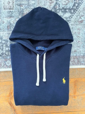 Mörkblå Polo Ralph Lauren Hoodie - Storlek L - Hej! 👋 Säljer en riktigt snygg mörkblå Polo Ralph Lauren hoodie i storlek L med gul pony-logga. 🐎 Tröjan är i mycket gott skick, välvårdad och helt utan hål eller fläckar. Självklart 100% äkta med originaltvättlappar kvar (ingen QR-kod på just denna årsmodell). Ett perfekt premium-basplagg som ger en skön stil till vardags! Fraktar snabbt! 3💬 100% 🚛📦✨ 