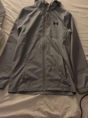 Grå vindjacka från Under Armour - Snygg grå vindjacka från Under Armour med huva och hel dragkedja framtill. Jackan har två sidofickor och en minimalistisk design med logga på bröstet. Perfekt för blåsiga dagar och enkel att matcha med olika outfits.