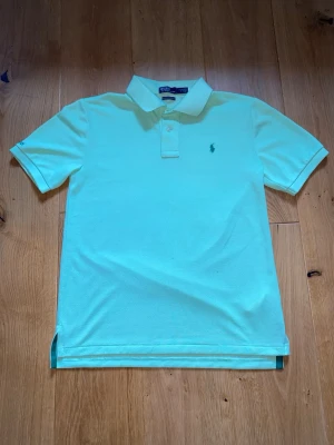 Ljus grön pikétröja från Ralph Lauren - Snygg ljusgrön pikétröja från Ralph Lauren med klassisk krage och korta ärmar. Tröjan har två knappar vid halsen och den ikoniska broderade loggan på bröstet. Perfekt för en avslappnad och stilren look.
