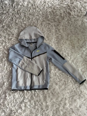 Nike ljusgblå hoodie med dragkedja - Nike hoodie i ljusblått med svarta detaljer och dragkedja framtill. Har huva, två fickor och en extra ficka med dragkedja på ärmen. Nike-logga i neongult på bröstet. Perfekt för en sportig och avslappnad stil.