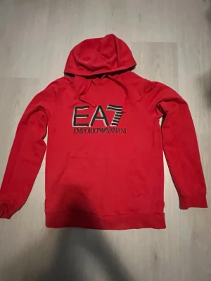 Röd hoodie från Emporio Armani EA7 - Snygg röd hoodie från Emporio Armani EA7 med svart och vitt tryck på bröstet. Klassisk huva med dragsko och långa ärmar. Perfekt för en avslappnad och trendig stil.