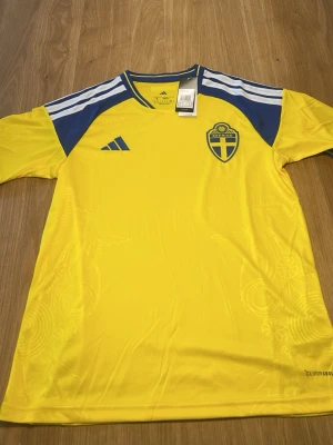 Svensk landslagströja Adidas gul - Gul svensk landslagströja från Adidas med blå detaljer och vita ränder på axlarna. Broderad landslagssköld på bröstet och Adidas-logga. Tillverkad i lätt och funktionellt material, perfekt för fotboll eller träning. 68 cm längd, 49 cm bredd 