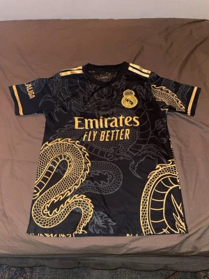  Kläder fotboll tröja i Madrid  - Svart Real Madrid matchtröja med guldiga drakmotiv och detaljer. Tröjan har korta ärmar, klubbmärke och Adidas-logga på bröstet samt 'Emirates Fly Better' tryckt fram. På ryggen står det 'Ronaldo' och nummer 7 i guld. Perfekt för fotbollsfans!