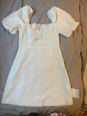 Vit puffärmad klänning Nelly x Glamorous - Superfin vit klänning från Nelly x Glamorous med puffärmar och rak passform. Klänningen har en söt, enkel design och är perfekt för sommardagar. Materialet känns lätt och luftigt, vilket gör den bekväm att bära. Styla med sneakers eller sandaler för en trendig look.