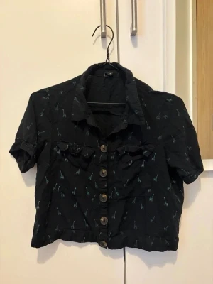 Svart kortärmad blus med knappar - Svart blus från Y.F.K. med korta ärmar och knäppning framtill. Blusen har små ljusblå giraffmönster och dekorativa volanger vid bröstet. Klassisk krage och en något croppad passform. Perfekt för en trendig och avslappnad look.