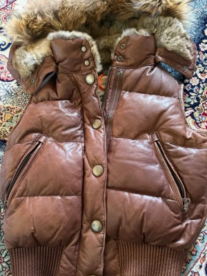 Brun dunväst med fuskpäls från Parajumpers - Snygg brun dunväst från Parajumpers med glansig yta och detaljer i fuskpäls på huvan. Västen har dragkedja, tryckknappar och två stora fickor med dragkedja. Ribbstickad mudd nedtill för extra komfort och värme.