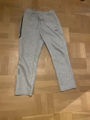 Grå Nocta mjukisbyxor från Nike - L - Snygga grå mjukisbyxor från Nike med dragsko i midjan och elastisk resår. Byxorna har raka ben, diskret Nike-logga på låret och tryck på ena benet. Perfekta för chill eller träning. Tillverkade i mjukt och bekvämt material.