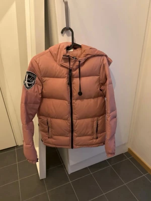 Rosa pufferjacka med huva - Säljer en snygg rosa pufferjacka med huva och svart dragkedja. Jackan har en stor patch på vänster ärm och två fickor framtill. Perfekt för kalla dagar och riktigt skön att bära. Jackan har en modern, boxig passform och dragsko i huvan.