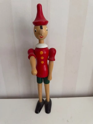 Pinocchio figur i trä Brio - Charmig Pinocchio figur i trä med röd hatt, röd tröja med gula knappar, gröna shorts och svarta skor. Figurerad med klassiskt långt näsa och målade detaljer. Perfekt som dekoration eller samlarobjekt.