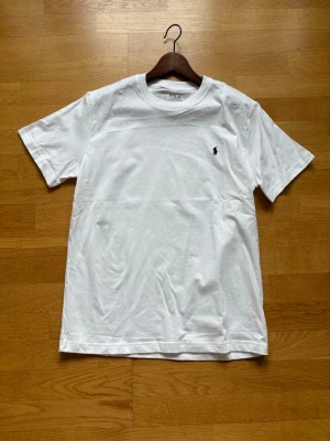 Vit t-shirt från Polo Ralph Lauren - Klassisk vit t-shirt från Polo Ralph Lauren med svart broderad logga på bröstet. Modellen är kortärmad och har en rund halsringning. Slim fit-passform och tillverkad i mjuk bomull för en skön känsla.