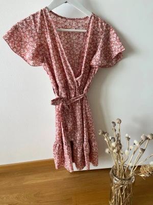 Rosa blommig omlottklänning med volang - Söt rosa kortklänning med små vita och röda blommor, omlottmodell och knytband i midjan. Klänningen har volangdetaljer och korta ärmar, perfekt för varma dagar. Tillverkad i mjuk bomull som känns skön mot huden.