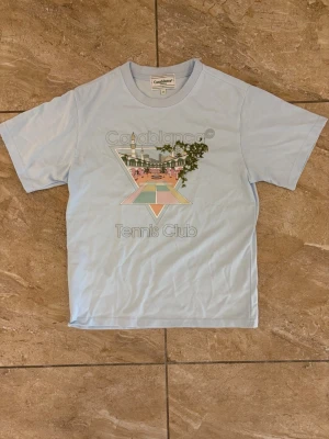 Ljusblå Casablanca Tennis Club t-shirt - Snygg ljusblå t-shirt från Casablanca med färgglad grafik och texten 'Tennis Club' på bröstet. Klassisk rund hals och korta ärmar. Perfekt för dig som gillar streetwear och vill sticka ut med en unik print. Tillverkad i mjuk bomull för skön känsla.